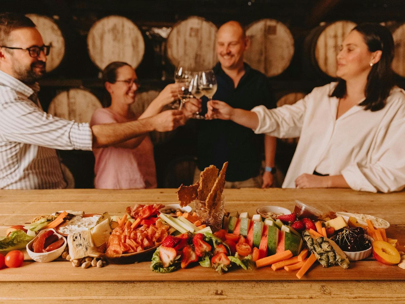 People enjoying a glass of wine and a massive cheese board in front of wine barrels