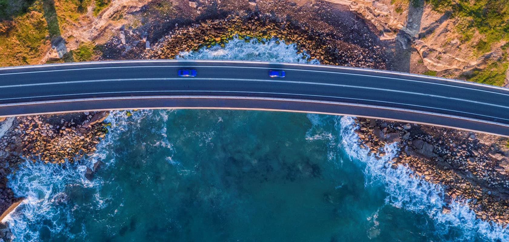 Birds eye of a highway over the edge of the coast with water underneath with two blue cars driving on the road
